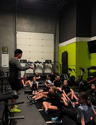 A coach instructs a group of seated athletes in a gym.
